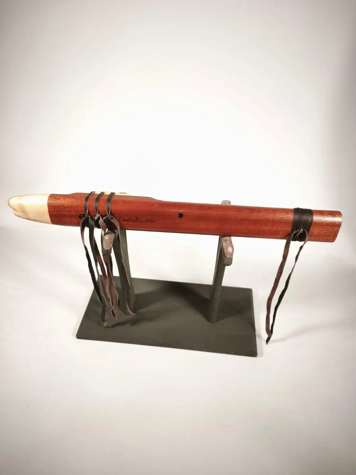 Reddish-brown sapele drone flute with a pale maple mouthpiece on a black stand, showing the back with the thumb hole, the ChantingDroneFlutes name burned into the wood and leather ties